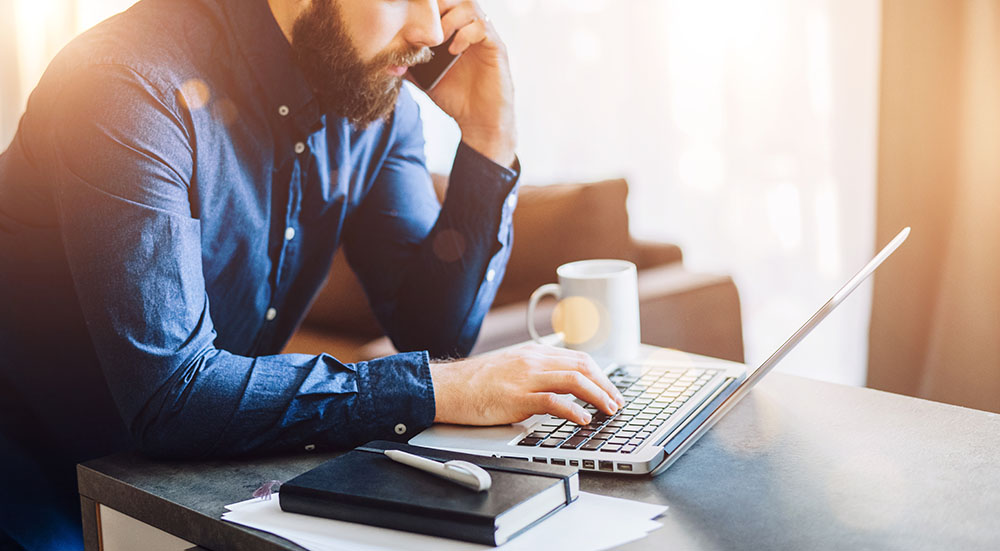 Young bearded businessman is sitting at table, working on computer, talking on cell phone. On desk notebook, documents.